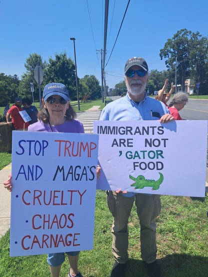 A woman protester is holding a sign that reads, "Stop Trump And Maga's Cruelty, Chaos and Carnage." Standing next to her is a man holding a sign that reads, "Immigrants Are Not 'Gator Food."