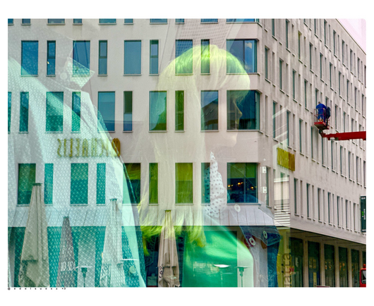 Farbfotografie: sich spiegelndes Schaufenster, dahinter schwach zu erkennen: zwei Schaufensterfiguren, von der einen nur der Oberkörper mit Hemd, von der anderen grünlich blonde lange Haare, einer weiblichen Figur mit Bluse, in der Scheibe spiegelt sich ein großes eckiges Gebäude mit vielen Fenstern, diese werden mit einem roten Hubsteiger gerade seitlich gereinigt