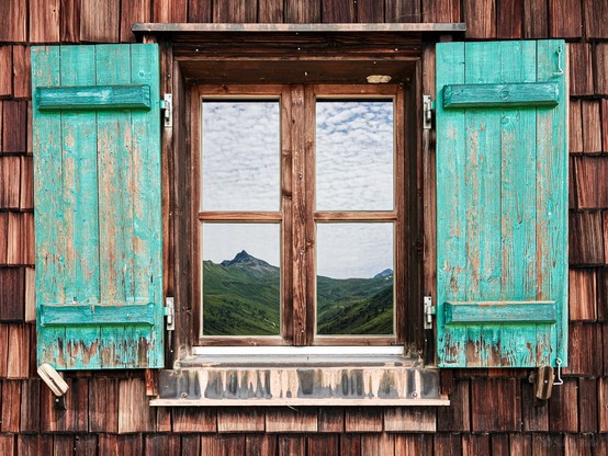 A wooden window with turquoise shutters. The shutters are  open, revealing a reflection of distant peaks under a partly cloudy sky. The window frame and the surrounding wooden wall feature a rustic design.