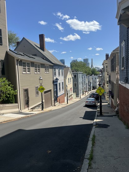 A row of various early 19th homes down a narrow descending street. There is a blue sky and a few small white clouds