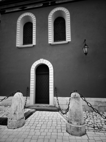 The image is a monochrome, eye-level, full shot of a building facade featuring a central arched doorway flanked by two prominent stone bollards connected by heavy chains in the foreground. Above the doorway are two arched windows with decorative grilles. A single, ornate lantern hangs on the wall to the right of the windows. The texture of the paved ground is visible in the foreground, leading up to the doorway. The overall impression is one of architectural detail and a slight sense of enclosure due to the chains and bollards. The two arched windows above the door, with their dark grilles, give the subtle impression of two eyes, with the door below resembling a mouth, creating a pareidolic face on the building.
