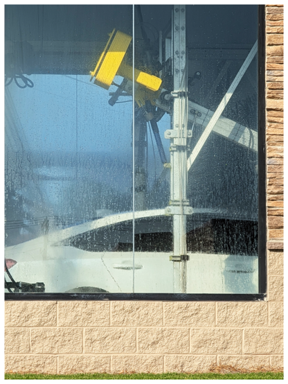 A white car is partially visible through a large glass window covered in water droplets. A yellow, mechanical car wash arm is cleaning the car. The window frame and a beige brick wall are at the bottom, with a small patch of grass outside.