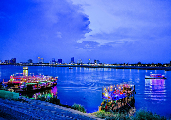 Photo shows two larger double floors restaurant boats on a river with lights and guards ready for an evening cruise on the river. The light is deep blue just after sunset. In the background there is the other river brim, with smaller lower houses and some larger high rises. In the north background the sky is divided in two. To the right it is light blue with few clouds lit up yellow from the sunset. To the left there are dark, stormy/rainy clouds. One of the clouds can seem to be forming an angry man’s profile. There are high expectations for a rainy night.