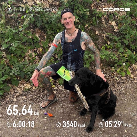 Das Bild zeigt einen tätowierten Mann (also ich), der kniet und einen schwarzen Labrador (Otto) hält. Der Mann trägt ein Laufshirt und scheint erschöpft, aber zufrieden zu sein. Die Zahlen im unteren Bereich deuten auf die Distanz (65.64 km), die Dauer (6:00:18), die verbrannten Kalorien (3540 kcal) und das Tempo (05'29"/km) hin.  Das Foto wirkt wie ein Schnappschuss nach einem langen Lauf im Wald.  Der Gesamteindruck ist einer von Ausdauer, Naturverbundenheit und der Freude an körperlicher Herausforderung, geteilt mit einem treuen Begleiter.