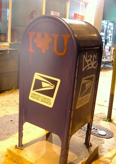 A blue Postal Service mailbox on a city sidewalk at night, lit by yellowish light. There is a graffiti tag on the front, and the side is spray painted with a red design. It looks like I Heart You, but the heart shape is replaced by a jigsaw puzzle piece.