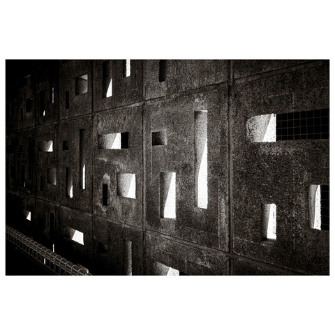 This black-and-white photo shows a concrete wall made up of modular panels, each pierced with a unique arrangement of geometric cutouts—rectangles, squares, and narrow slits. Sunlight streams through from the other side, casting abstract, high-contrast shapes and giving the scene an almost architectural-art-installation feel. Despite its complexity, the setting is mundane: this textured wall belongs to a paid parking lot in Nagoya.