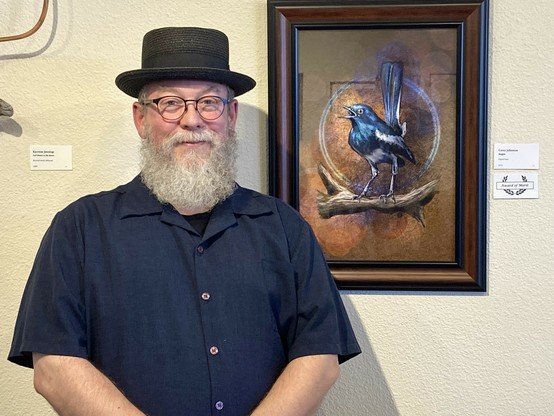 PHotograph of an artist with a beard in a pork pie had standing next to a digital art piece in a frame of an illustrated Magpie in a gallery.