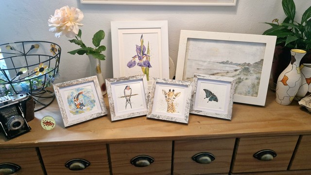 Auf einer Holzkommode stehen insgesamt 6 handgemalte Bilder, 4 kleinere quadratische Tier Illustrationen jeweils mit dem Motiv: Koi Fisch,  Schwalbe ,  grüner schwalbenschwanz-schmetterling, Giraffe,   und ein größeres Bild mit einer illustrierten Schwertlilie, und dahinter  eine unverkäufliche landschaftszeichnung der SchleiGegend.