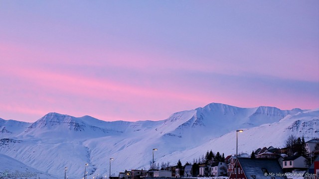 A colour photo of a landscape mostly filled by the sky. The foreground is a small street of houses with snow-topped roofs, with a row of lit streetlights and trees beyond them. Behind these is a line of majestic mountains, flanks heavy with snow and ice, their sun-ward faces slightly lit. The sky above is streaked with sunrise colours, pink and streaky against cyan.