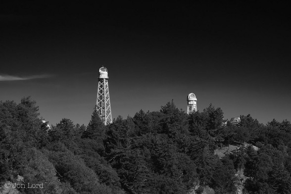 This is a high contrast black and white landscape photo of two white solar telescopes mounted on tall towers. In the foreground is dense, tree covered rising terrain reaching a peak. There are few breaks in the forrest. On the peak are two bright white towers rising high above the trees. The tower to the left of centre is the taller and consists of a lattice construction with a small observatory on top with its lid open and facing the camera. To the right of centre and a little more distant is a shorter tower similar to the first. There is a building or two further to the right but partially obscured by the forrest. Above is a near clear and cloudless sky, just a wisp of cirrus high cloud to the far left. Above a jet black sky. 
The location: The Mount Wilson Solar Observatory, 1700 metres above Pasadena in the San Gabriel Mountains, California (2011).