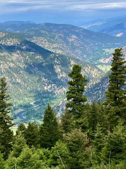 The view looks down a steep slope and over trees far below at a narrow valley where a stream meanders. All around are wooded mountains, the clouds turning them light and dark in unusual places. The sky is mostly cloudy.
