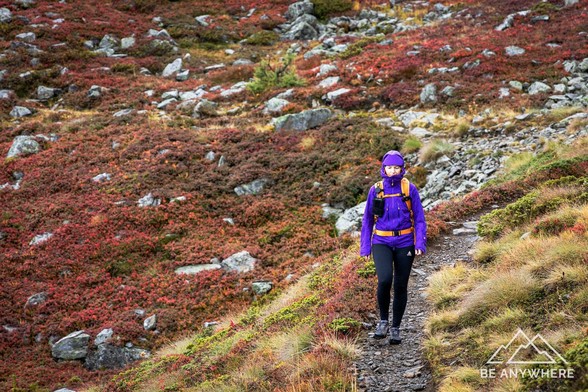Wanderin in Regenkleidung auf einem schmalen Bergpfad, umgeben von herbstlich verfärbtem, nassem Heideland und Felsblöcken. Die Stimmung ist ruhig und zurückgezogen.