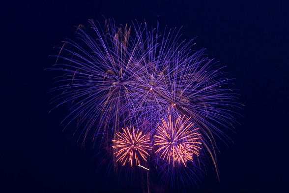 Big purple fireworks explode as there main subject two smaller red fireworks explode closer to the ground against a clear dark blueish purple sky.