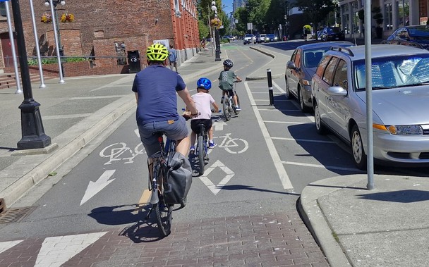 Two small children riding bicycles in front of an adult on a bicycle, in a two-way protected bike lanes.