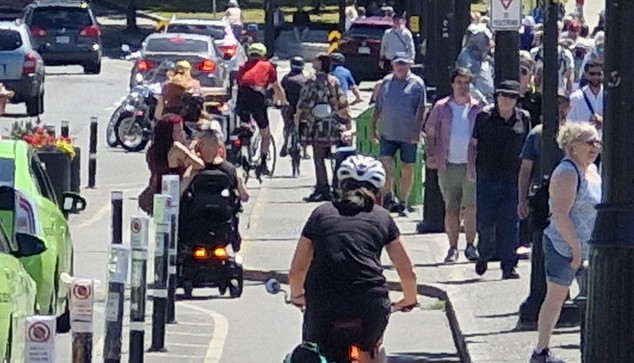 Wheelchair with two people on it on busy protected bike & roll lane beside busy sidewalk.