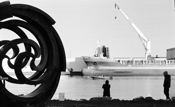 Fishermen, Fox River, Green Bay, May 2025. Black and white photo, you can see the silhouettes of two fishermen and a public sculpture. In the background there is a freighter moving things into a warehouse facility. The Fox River is a working river that gets lake freighter traffic from all over, occasionally we get international ships too.