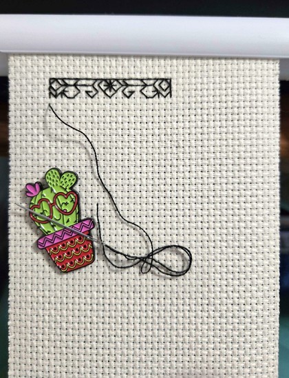 a white piece of Aida cloth is stretched on a frame. Three rows of a blackwork embroidery patter have been started, and the thread and needle is attached to a magnetic needle holder in the shape of a cactus in a pot with heart shaped glasses and a smile.