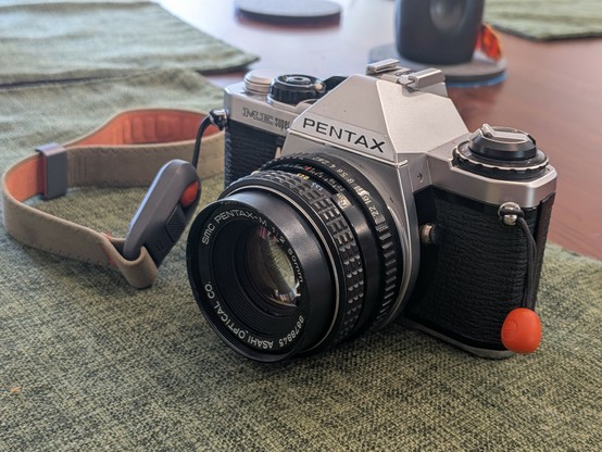 My Pentax ME Super with a sage green suede wrist strap. It is sitting on a sage green place mat on my walnut dining room table.
