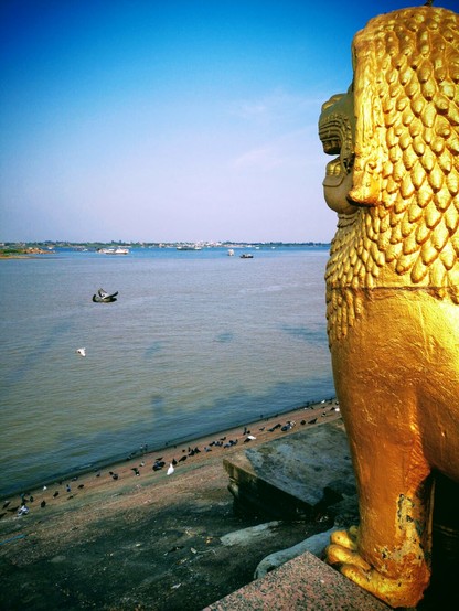 Photo shows a busy river meeting point, filled with smaller and larger boats and ferries crossing and going up and down the rivers. In the background is the northern river brim, with some small houses but most green bushes and trees. To the right in the front there is a statue of a golden lion guardian statue in the style of Khmer art. it is looking out over the river. To the left there are two pigeons flying by, sharing the lion statue’s view. It is a sunny day, and the sky have some very lightly clouds.