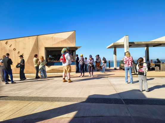 A beachside kiosk surrounded by people casually waiting for their orders. It's a sunny day and the sky above is a cloudless, deep blue. Glimpses of the ocean can be seen beyond the kiosk handrail.