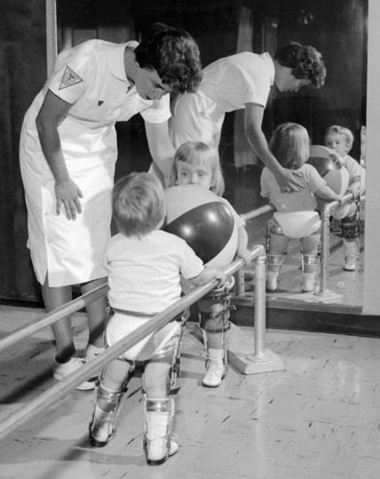 This is a black and white photograph depicting a therapy session for children affected by polio.

In the foreground, two young children are shown from behind, both wearing metal leg braces extending from their hips down to their feet. They are holding onto parallel bars, which are common equipment for gait training and physical therapy. 

The child on the right is holding a large, two-toned (likely black and white or dark and light) inflatable ball in front of them, using it to lean against the parallel bars.

Behind the two children, a woman in a white uniform, resembling a nurse or therapist, is bending over and assisting them. She has short, dark hair and is wearing a white dress or uniform with short sleeves, and a patch (possibly a Red Cross or medical symbol) on her right sleeve. Her expression is focused and caring as she looks at the children.

To the right side of the image, a large mirror reflects the scene. In the reflection, we can see the front of the woman assisting, and the back of the child on the right, still holding the ball and wearing braces. Another child's reflection is also visible in the mirror, slightly behind and to the right of the first two, also wearing braces and looking towards the ball.

The overall tone of the image is one of medical care, determination, and the challenges faced by children undergoing physical rehabilitation.