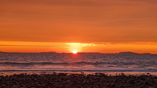 Historical Documentation in the form of a photograph of a rising sun over the ocean bay at Yirrell Beach in Winthrop, MA. The sky is filled with vibrant orange and yellow hues, with the sun partially visible in the center of the image, just behind a distant island. The sun is rising over the horizon, casting a warm glow over the water and the horizon as it sets the sky a blaze in a gradient of orange hues. The water is calm as the waves gently crash onto the shore. In the foreground, there is a rocky beach with small pebbles scattered across it. On the right side of the image, there are a few small islands visible. The overall mood of the photograph is peaceful and serene as we witness the dawn of a new day.