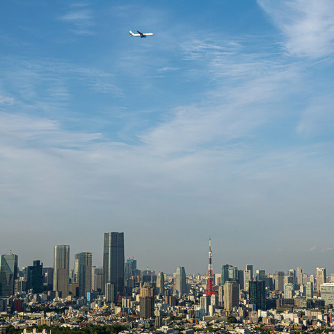 Skyline view of Tokyo, with Tokyo Tower and the Azabudai Hills Tower. Far above in the sky, a JAL airliner goes by.