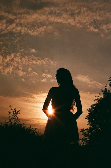 Colour film photo of a long-haired young woman silhouetted against a golden sky with a few clouds and the setting sun, which makes a sunstar between her arm and hip