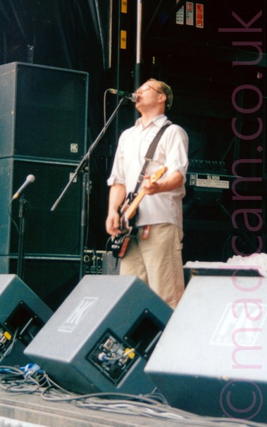 A man with short brown hair, wearing a white shirt and khaki pants, playing a black electric guitar and singing into a microphone on a stand in front of him on an open air stage.
The mic stand is obviously set up for someone a little bit taller than him!
Monitor speakers litter the front of the stage, with vertical stacks of speakers and amps in the background.