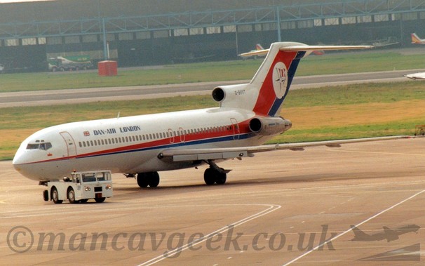Side view of a 3 engined jet airliner with the engines mounted on the rear fuselage, being towed from right to left, turning back to the right again.
The plane is mostly white, with a thin red and blue stripe running along the body, getting wider as it moves rearwards before sweeping up in to the tail.
There is blue "Dan-Air London" text on the upper forward fuselage, and the black registration "G-BHVT" on the side of the centre intake above the rear fuselage.
There is a white circle on the tail, containing a 4-pointed compass star with a red and white barred flag behind.
The plane is being towed by a low-slung white truck turning towards the right of the screen.
Grey concrete apron fills the foreground, with grass either side of a runway in the background leading up to a wide black hangar in the distance.