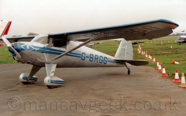 Side view of a high-winged single-engined light aircraft parked on a tarmac apron facing to the left.
The plane is mostly in a bare metal finish, with a pair of blue stripes running along the body, getting narrower as they move to the rear, overlaid  with the blue registration "G-BRGG" on the rear fuselage, which is repeated under the left wing.
Another pair of blue stripes adorn the stripes on the spats over the main wheels.
Struts support the wings, connected to the body just above the main gear legs.
Grey tarmac apron fills the foreground, with a row of orange and white traffic cones separating that from muddy grass on the right of the frame.
There are more light aircraft parked on grass in the background, under cloudy grey sky.