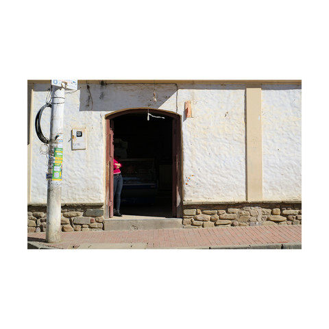 Photo of a woman in red sweater standing at the door of her shop, hiding her face in the shadow, in San Lorenzo, Tarija, Bolivia.