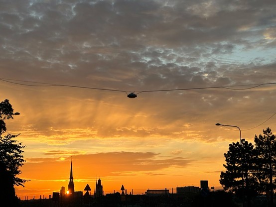 the setting sun peering out from behind the silhouettes of distant buildings, including a church spire. The bottom of the picture is in shadow, and the sides are the silhouettes of trees, and a golden sunset illuminating banks of clouds fills the rest. An overhead street lamp on a cable runs across the photo just above halfway up.