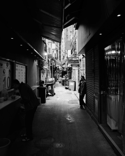The image shows a narrow Hong Kong alley in black and white. It features dim lighting, with a person standing and leaning against a wall. The alley is lined with old buildings and strewn cables. There are a few people going about their activities, adding life to the scene.