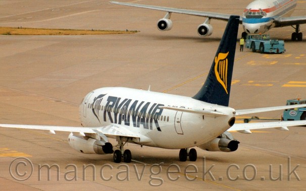 Rear side view of a twin engined jet airliner taxiing from right to left.
The plane is mostly white, with large, blue, billboard-style "RyanAior" titles next to a blue, winged Irish harp.
The tail is the same blue, with a larger, yellow, winged Irish harp.
Grey concrete apron fills the frame, with a white, 4 engined jet airliner facing the camera, being pushed backwards by a blue tug in the top right corner, it's right wing stretching across the top of the frame.