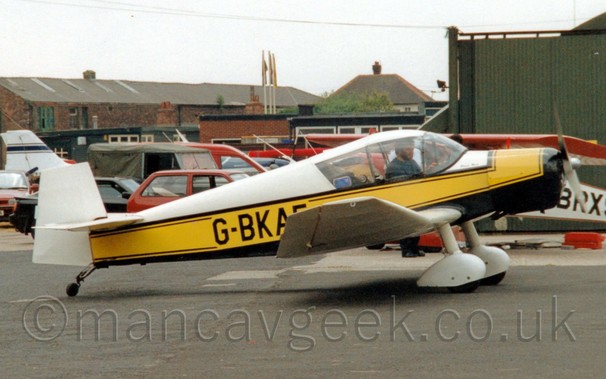 Side view of a single engined light aircraft parked on tarmac facing to the right.
The plane is mostly white, with a black belly, and a thick yellow stripe running along the body, over-laid with the black registration "G-BKAE" on the rear fuselage, the last letter partially obscured by the slightly up-turned wing-tip.
White spats cover the fixed main landing gear, under the wings.
A man with short dark hair and beard, wearing a blue top, can be seen in the cockpit, possibly running an engine test, as the propellor on the nose looks to be slightly blurred.
Black tarmac apron fills the foreground, with a green hangar on the right of the frame in the background, with cars parked behind the plane on the left, with brown brick buildings beyond that.
Flat white sky fills the rest of the frame.