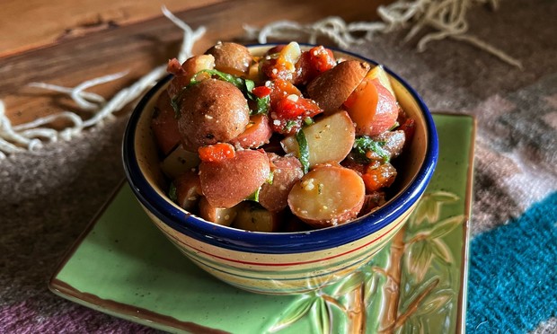 A green square plate with a bamboo motif with a small, round, colorfully painted bowl on top, both sitting on a woven runner on a wooden table. The bowl contains a mixture of diced baby red potatoes, bits of roasted red bell peppers, black and green olives, basil, and a seasoned dressing (olive oil, balsamic vinegar, garlic salt, black pepper, parmesan cheese, oregano, red pepper flakes).