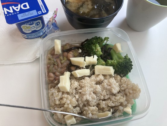 A meal in a clear container featuring brown rice topped with white powder, boiled beans, and pieces of broccoli. Nearby is a cup of miso soup and a container of yogurt, alongside a napkin.