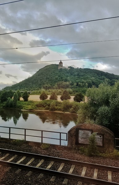 Vordergrund, Bahngleise. Dahinter: ein Fluß, die Weser. Dahinter: ein bewaldeter Berg, darauf das "Kaiser-Wilhelm-Denkmal" in Porta Westfalica. Das Denkmal hebt den rechten Arm hoch — zum Gruß. Wurde renoviert, damit Privilegierte dort ihre Hochzeiten feiern können. Graue Wolken darüber.