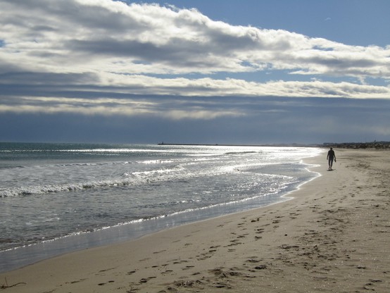 Ein Mensch läuft auf dem Sand am Meer entlang. Viele Spuren im Sand und der Himmel im Hintergrund ist etwas duster - ein guter Kontrast zu dem glitzernden Wasser.