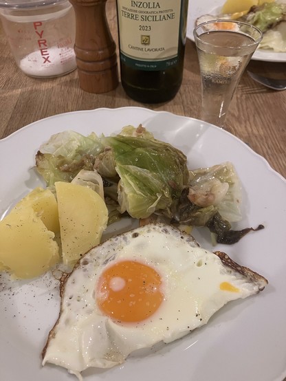 A plate with a fried egg featuring a runny yolk, boiled potatoes, and sautéed greens. In the background, there is a bottle of Inzolia wine and a shot glass.