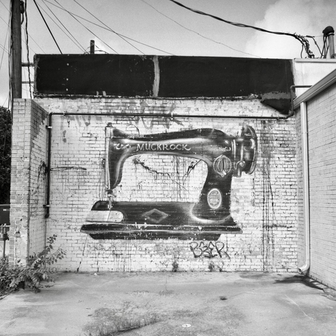 A black and white film photo. Graffiti art of a giant antique sewing machine is shown on an old white brick wall in Smithville, Texas. Sunday, June 29, 2025.