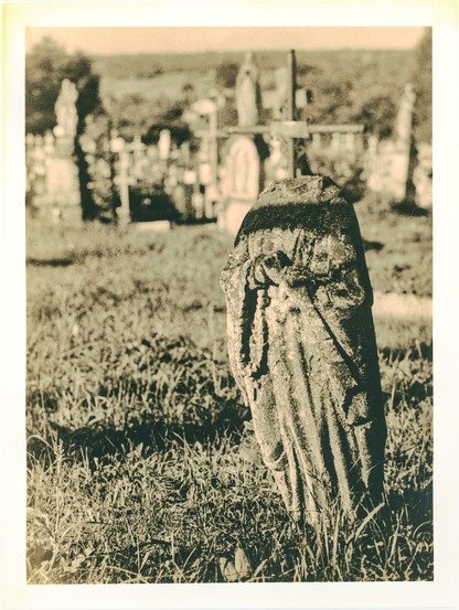 A weathered stone statue, likely of a saint or angel, stands alone in the foreground of a rural cemetery. The figure’s head is missing, and its surface is heavily eroded, covered with moss or lichen, as if it were silently crumbling under the weight of time. In the softly blurred background, crosses and gravestones stretch across the field under warm, low sunlight. The image was printed using the lith print technique, which enhances the dramatic tonal range and grainy texture, giving it an aged, almost haunted appearance.