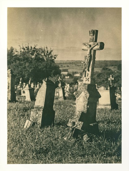Leaning and broken gravestones emerge from a hillside cemetery, surrounded by wild grass. The most prominent marker is a tall stone crucifix with a time-worn sculpture of Christ, still standing amid the decay. Soft afternoon light casts long shadows, while rolling hills fade into the horizon. The lith print process imbues the scene with a deep sense of melancholy, intensifying its gothic and timeless mood.