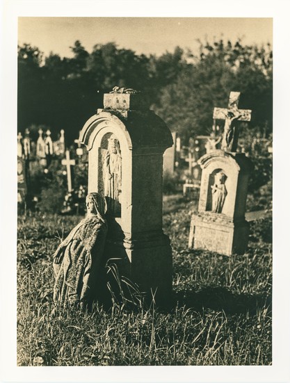 A statue of a mourning woman leans against an arched tombstone in a grassy field, her posture frozen in eternal sorrow. The surrounding gravestones, many crowned with crosses and effigies, recede into the background beneath the fading sunlight. This lith print captures the emotional weight and textural richness of the scene, evoking a quiet reverence and the haunting beauty of forgotten places