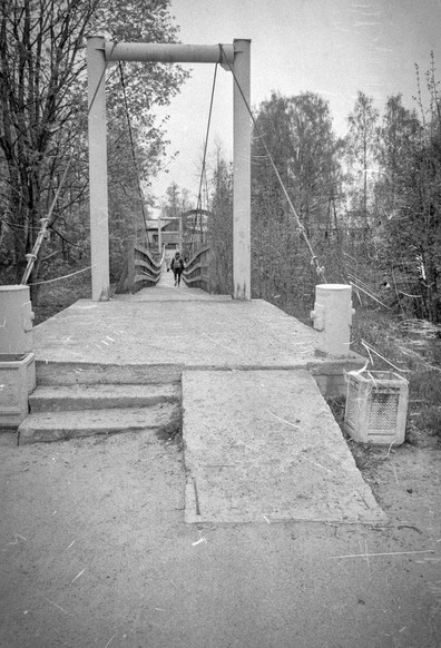 The black-and-white photo shows a suspension bridge. A person can be seen on the bridge. Trees grow on both sides of the bridge, creating an atmosphere of privacy and tranquility. The bridge's graceful lines contrast with the massive surrounding landscape.