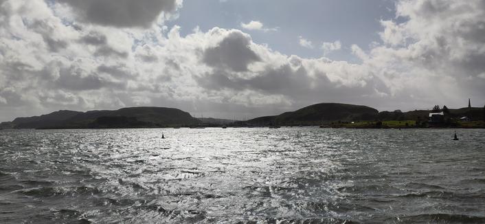 Blick von der Strandpromenade in Oban (Stadt in Schottland) Richtung Norden aufs aufgewühlte Meer. Zwischen Meer und Himmel ist die Insel Kerrera zu erkennen. Nach einem sehr stürmischen Vormittag mit viel Regen ist jetzt der Himmel am Nachmittag blau mit Wolken. Es ist weitestgehend trocken. Es weht immer noch ein starker Wind.