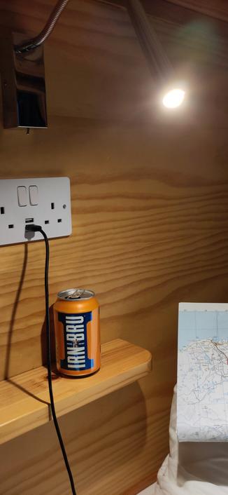 Photo einer Holzwand. Darin eingebaut eine typisch englische Steckdose mit Schaltern, hier zusätzlich mit zwei USB-A-Ladeports. Auf einem kleinen Regal darunter steht eine Flasche Irn Bru. Außerdem ist eine Straßenkarte mit dem Ausschnitt des nordöstlichen Ende Schottlands zu sehen.