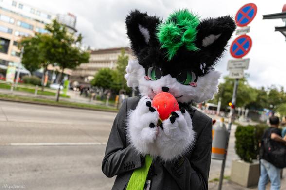 Vanj fursuit in a business suit with a green tie, tasting a strawberry outside, visible street, trees and buildings in the background.