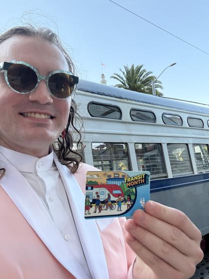 An enby wearing a pink suit and blue glasses holding a Transit Month Clipper card in front of a streetcar with grey and blue livery